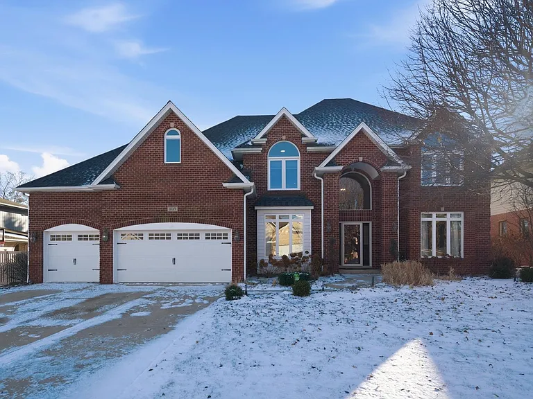 Large brick house with two-car garage, snow-covered yard, trees, and clear sky.
