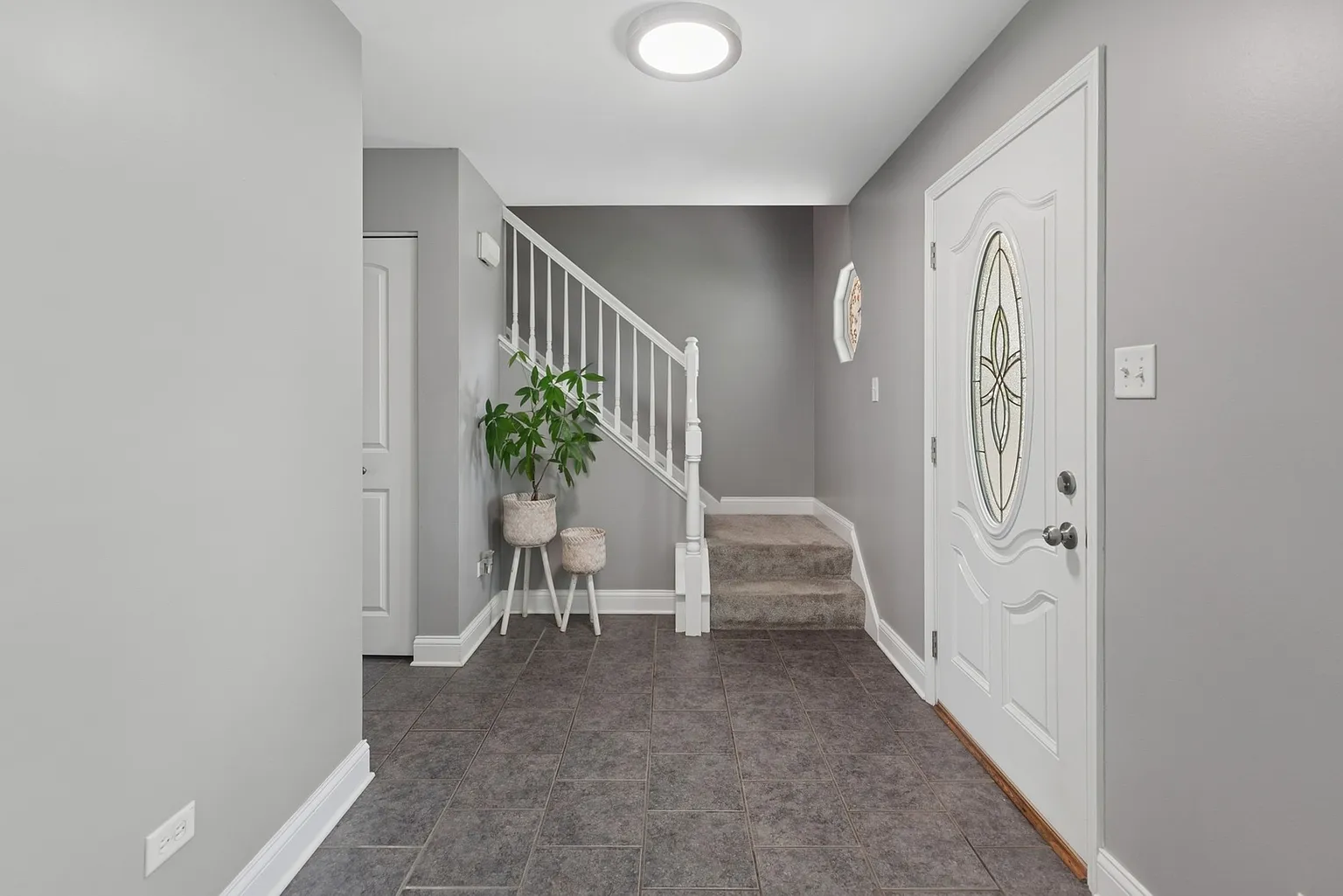 Modern entryway with staircase, potted plant, gray walls, and tiled floor.