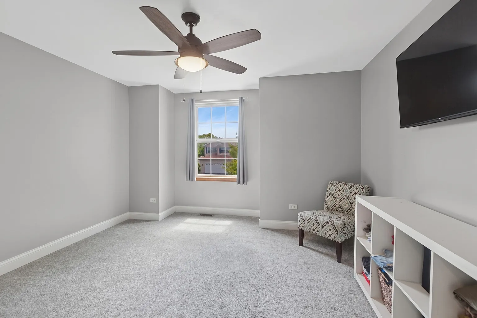 Well-lit bedroom with gray walls, carpet, ceiling fan, window, and upholstered chair.