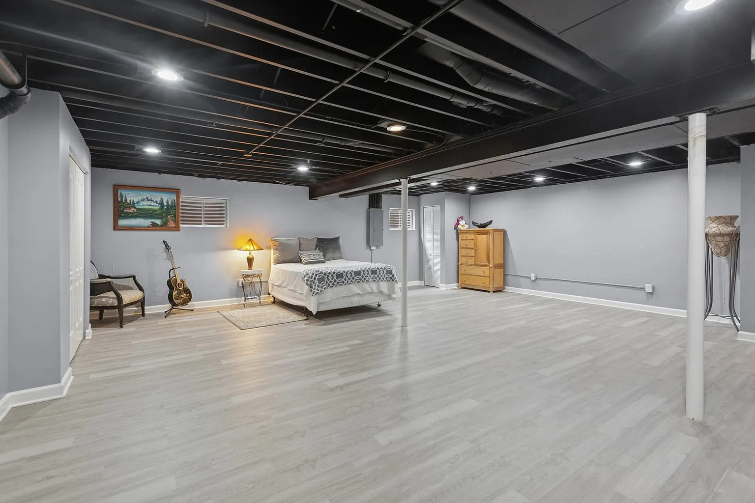 Finished basement with bed, wooden dresser, guitar, gray walls, and ceiling lights.