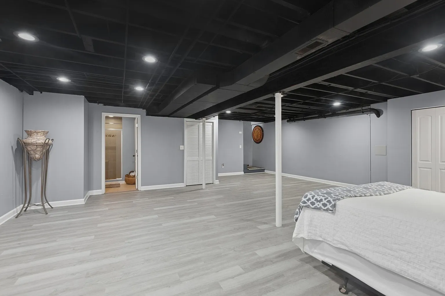 Spacious unfinished basement with gray walls, exposed beams, and white bed.