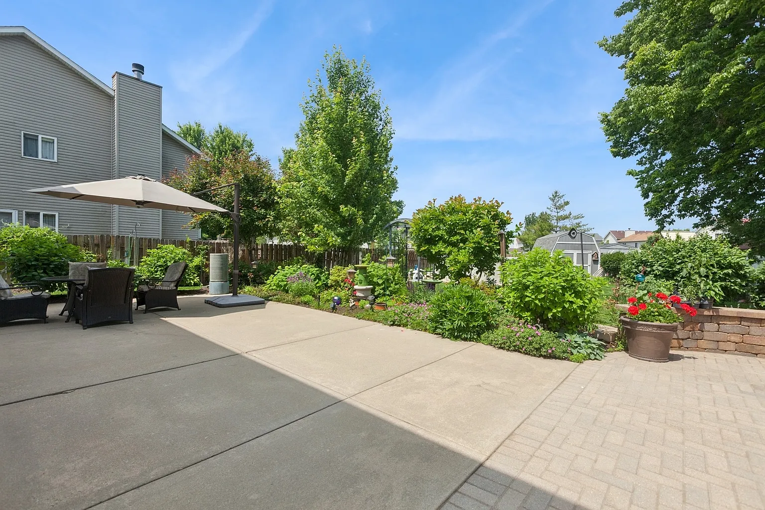 Backyard patio with table, umbrella, greenery, flowers, and small trees.