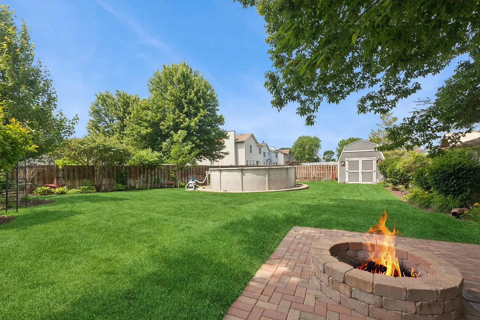 Spacious backyard with green lawn, above-ground pool, trees, and brick patio fire pit.