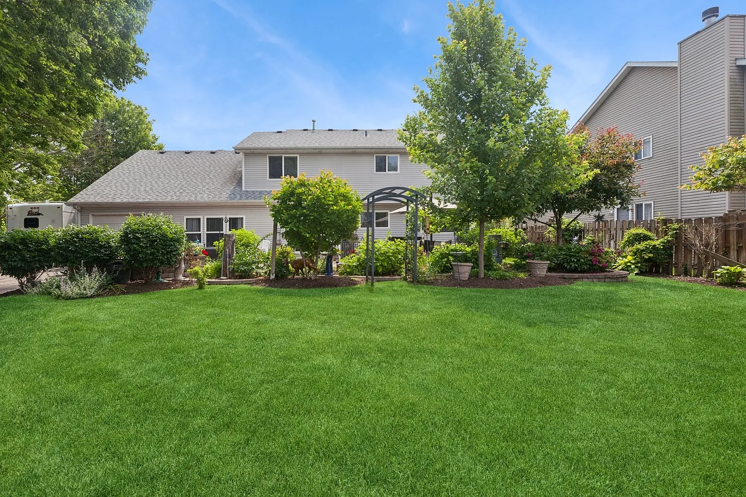 Well-maintained backyard with green grass, shrubs, trees, decorative plants, and residential house.