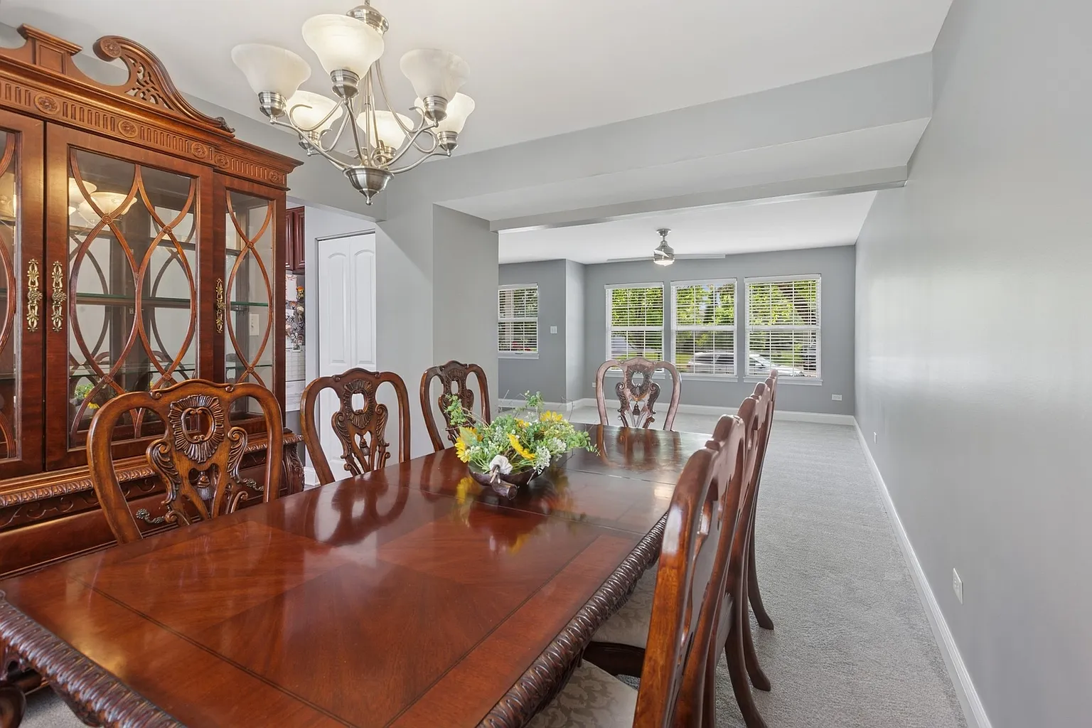 A dining room with a wooden dining table and chairs, a chandelier overhead, and a china cabinet against the wall. The walls are painted gray, and there are windows allowing natural light into the space.