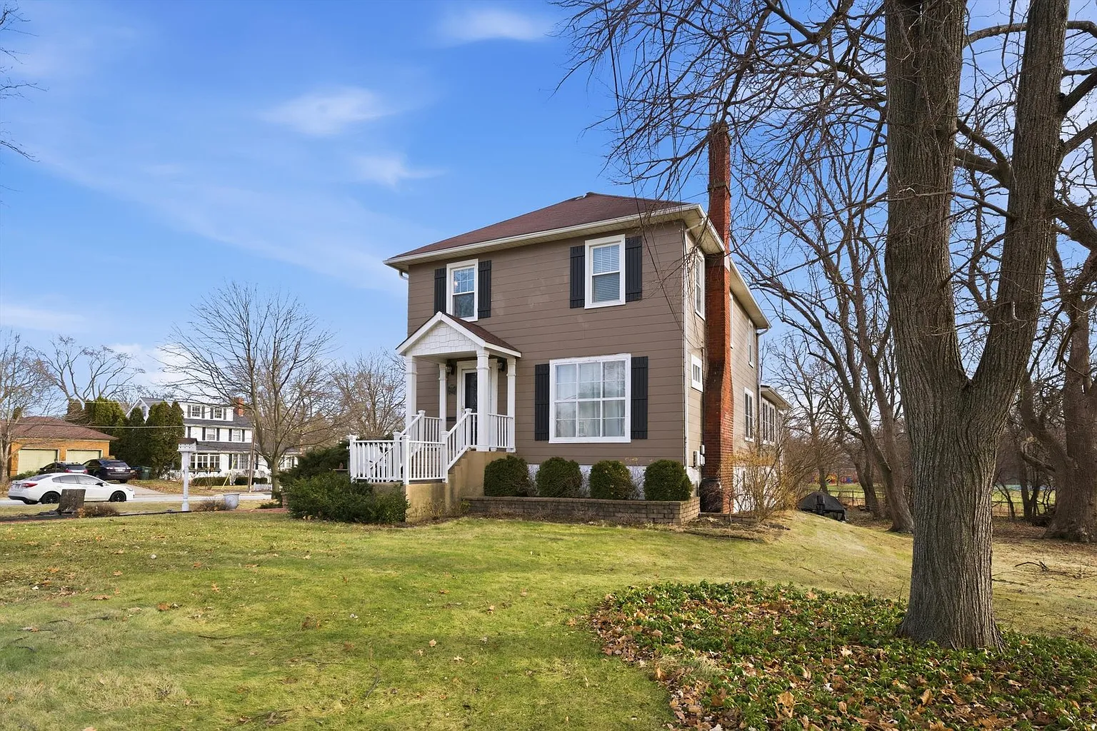 Two-story brown house with white trim, porch, lawn, hedges, and bare trees under blue sky.