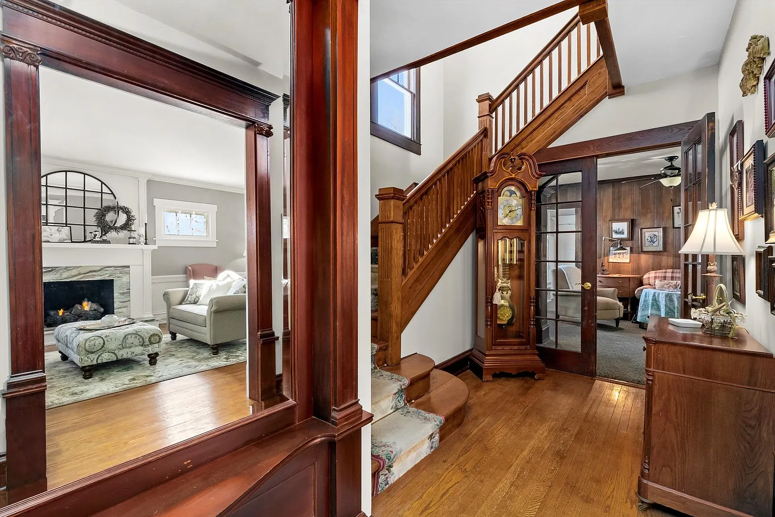 Hallway with wooden staircase, grand clock, and access to cozy living room with fireplace and seating.
