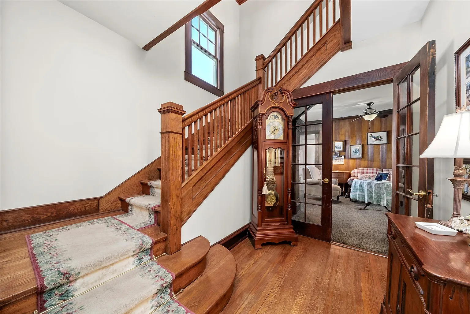 Wooden staircase with rug, grandfather clock, doorway, and neutral-toned living room.