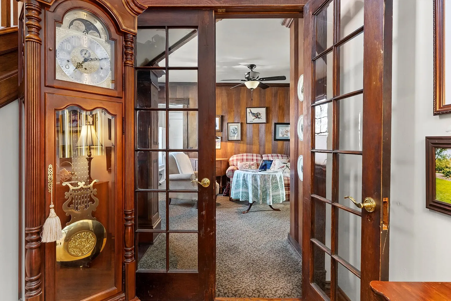 View through wooden double doors into cozy room with patterned sofa, ceiling fan, wall art, and grandfather clock.