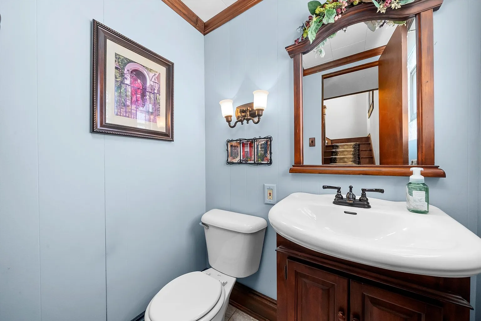 Small bathroom with toilet, sink cabinet, mirror, wall lights, light blue walls, and artwork.
