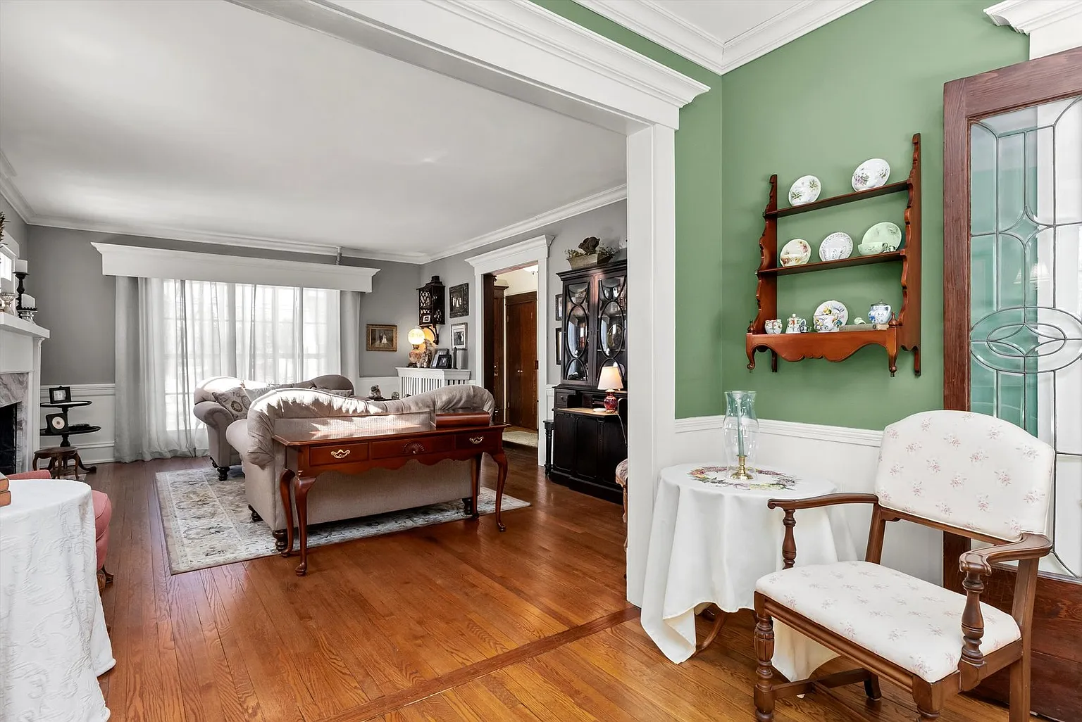Spacious living room with green accent wall, hardwood floors, large windows, elegant furniture, and shelves with decor.