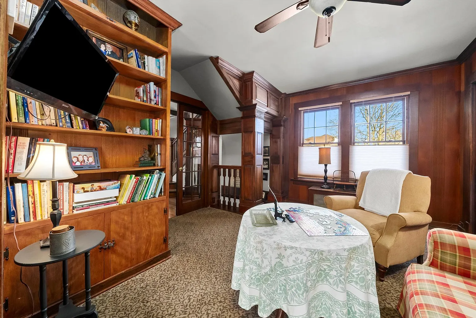 Cozy room with wood paneling, armchair, bookshelves, window light, and small table with lamp and supplies.