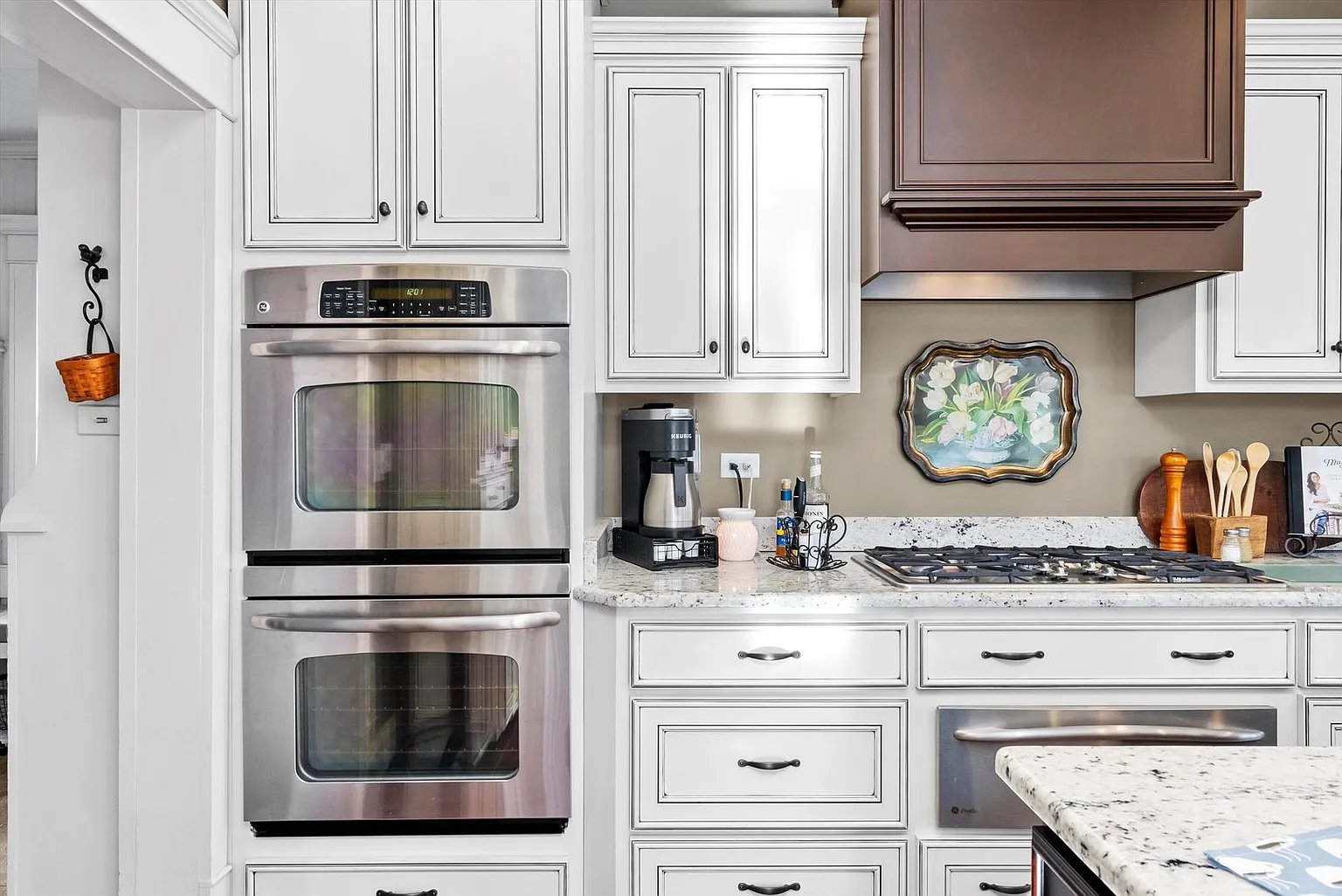 Modern kitchen with white cabinets, double ovens, coffee maker, utensils, and light stone countertops.