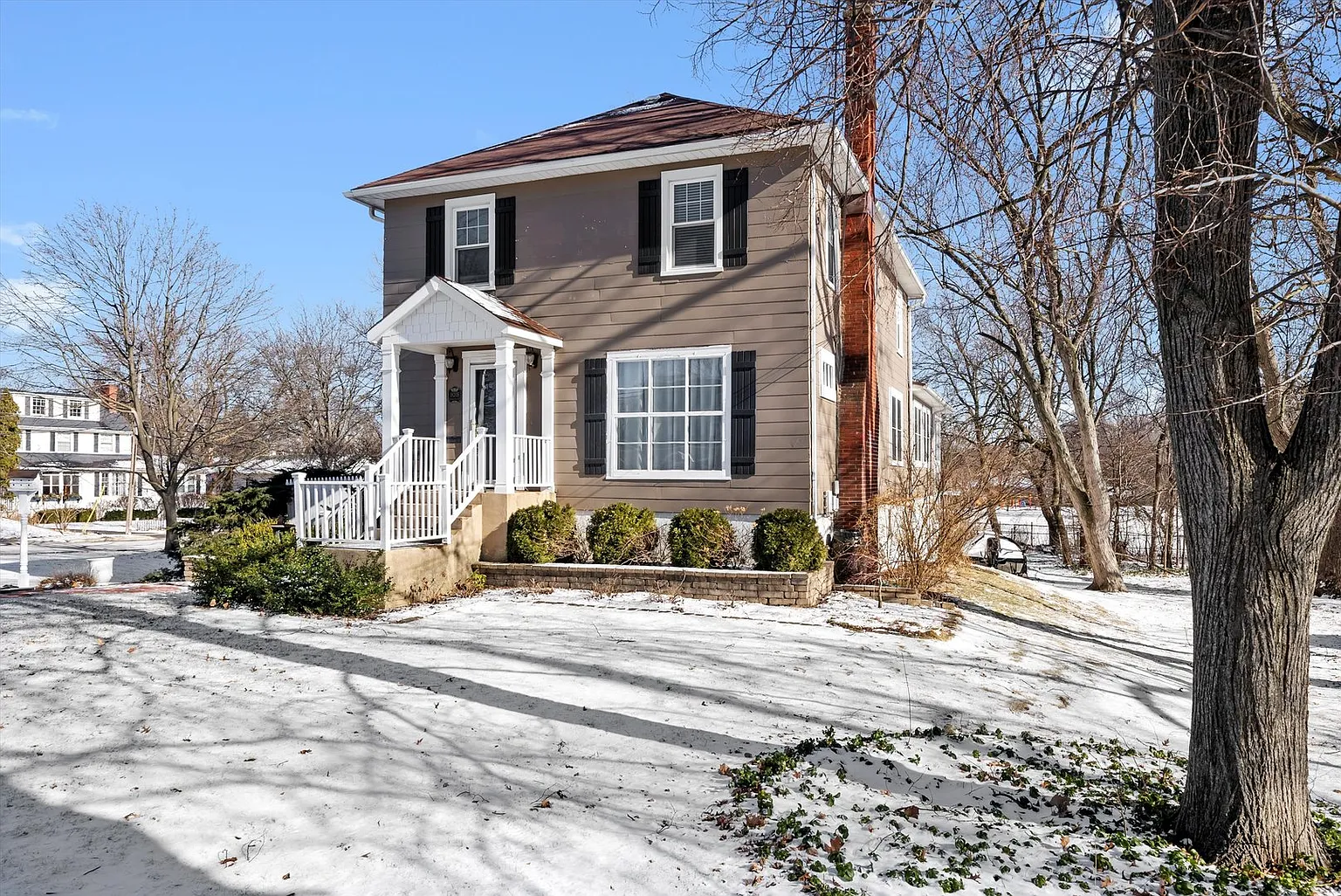 Two-story beige house with black shutters, snowy yard, bare trees, and blue sky.
