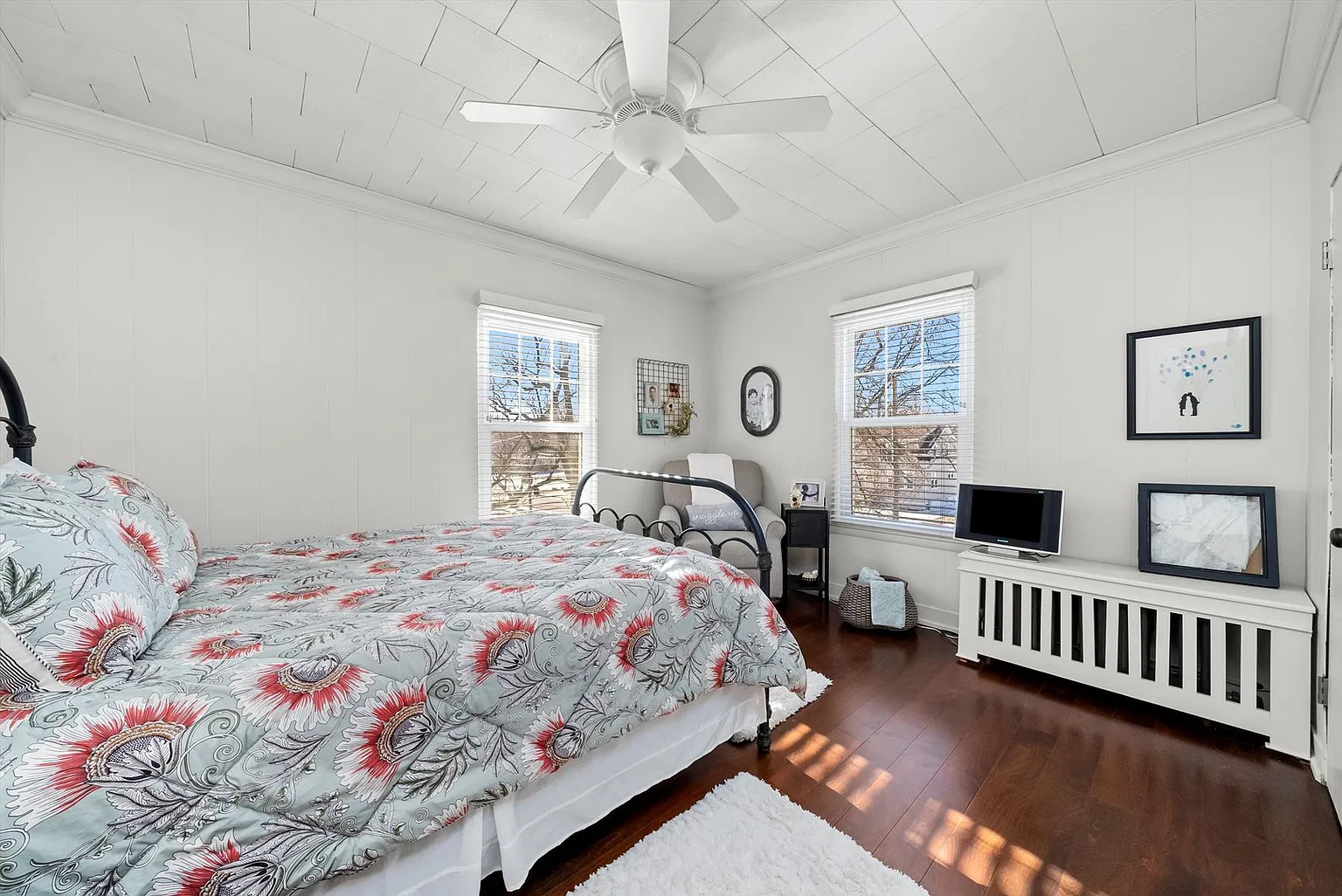 Well-lit bedroom with bed, floral comforter, windows, TV stand, and wood floors.
