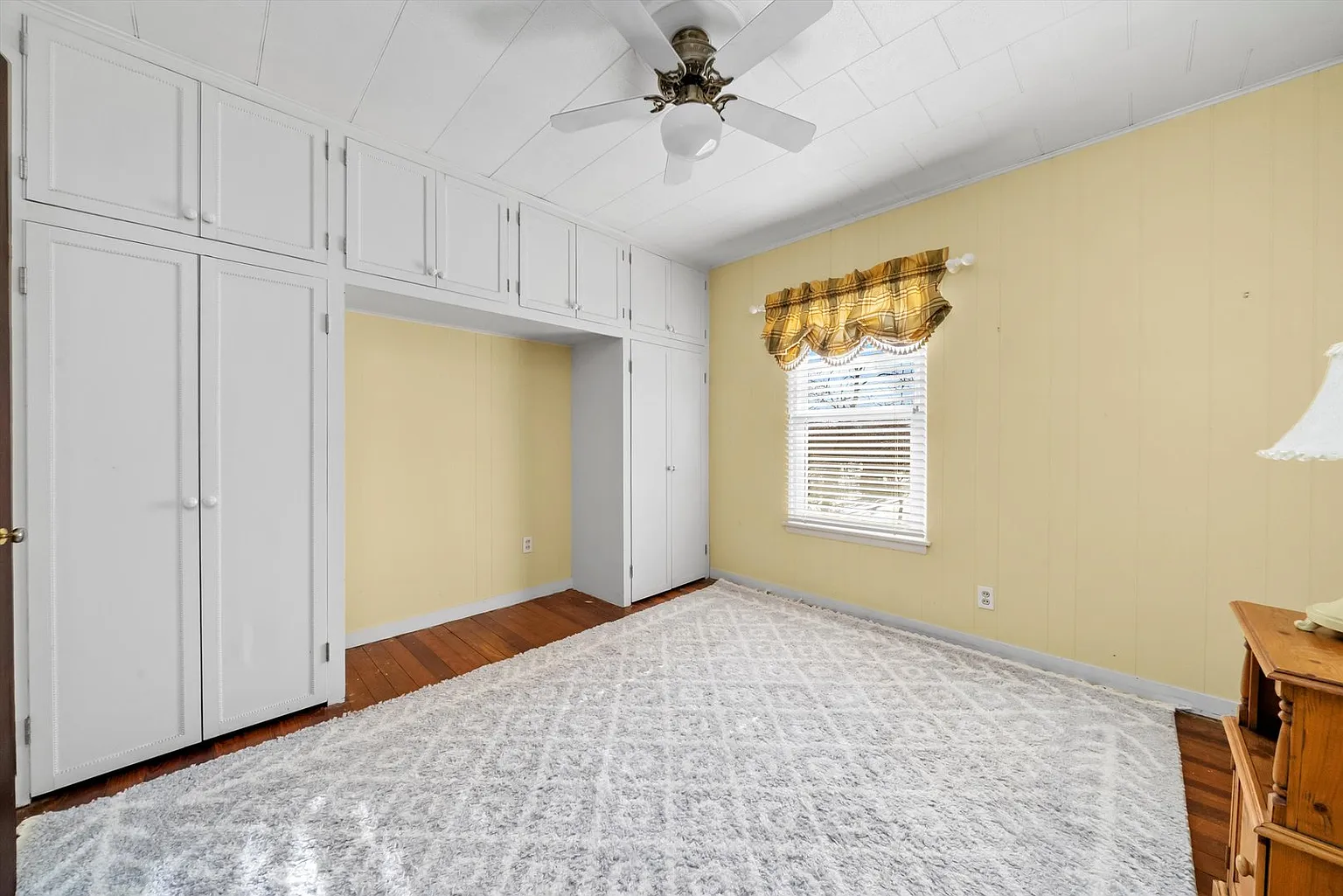 Bedroom with yellow walls, ceiling fan, hardwood floors, rug, cabinets, window, and small table.