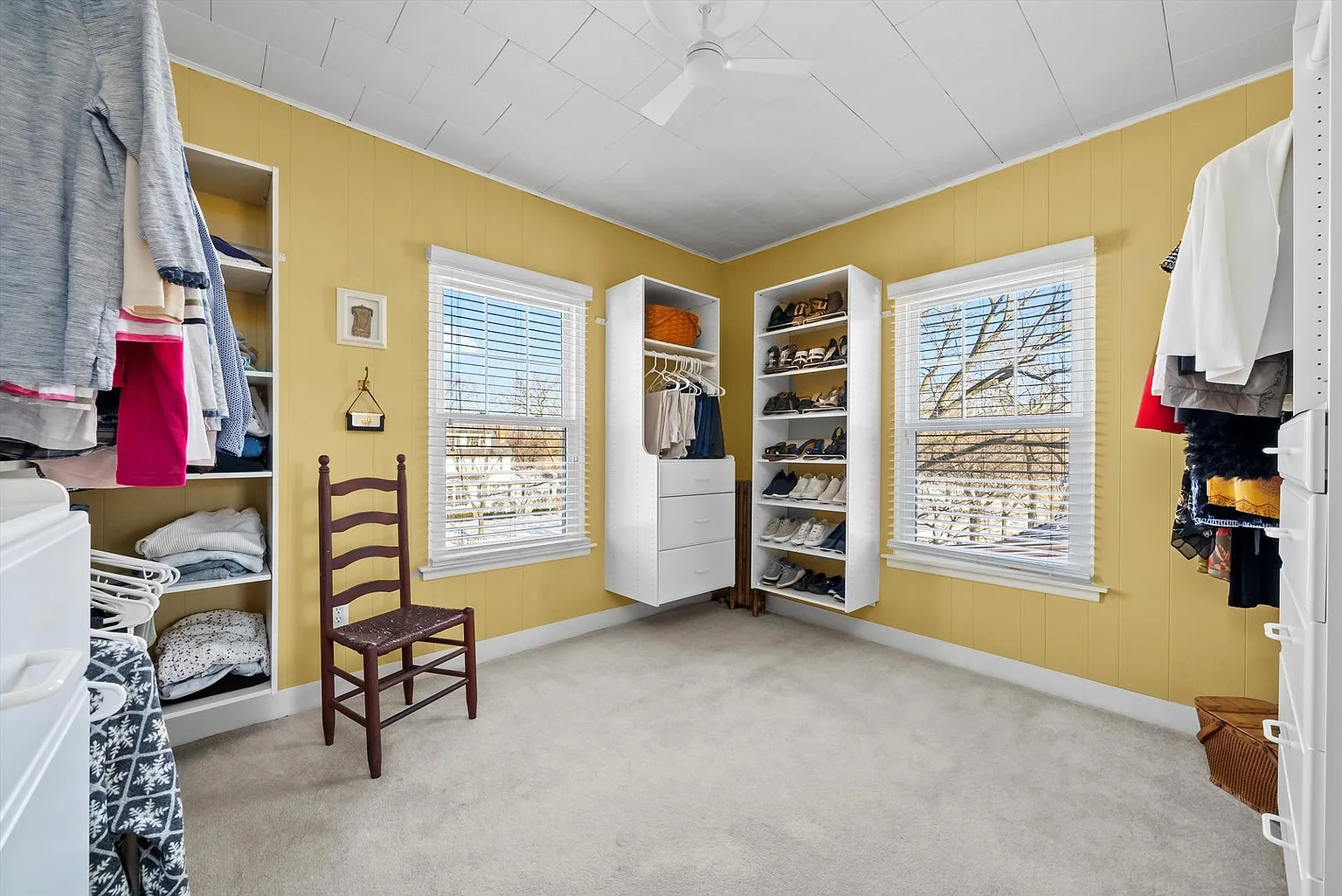 Spacious bedroom with yellow wall, windows, wooden chair, and organized closet.
