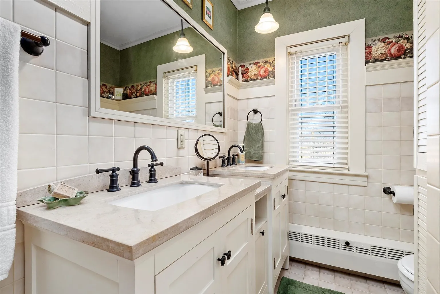 Bathroom with double vanity, two sinks, black faucets, large mirror, green walls, floral wallpaper, and natural light.