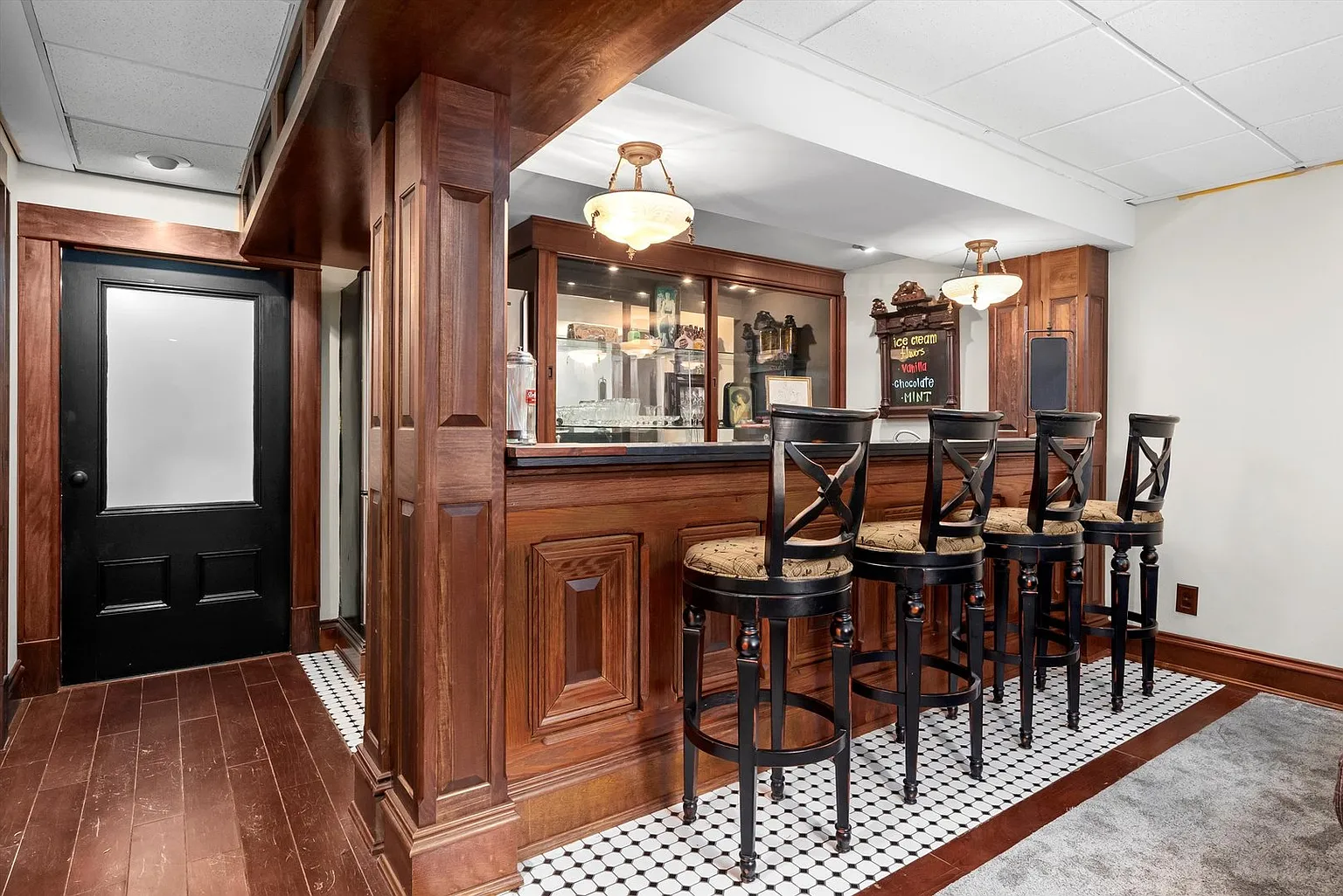 Wooden bar area with stools, glass cabinet, and black and white tile floor.