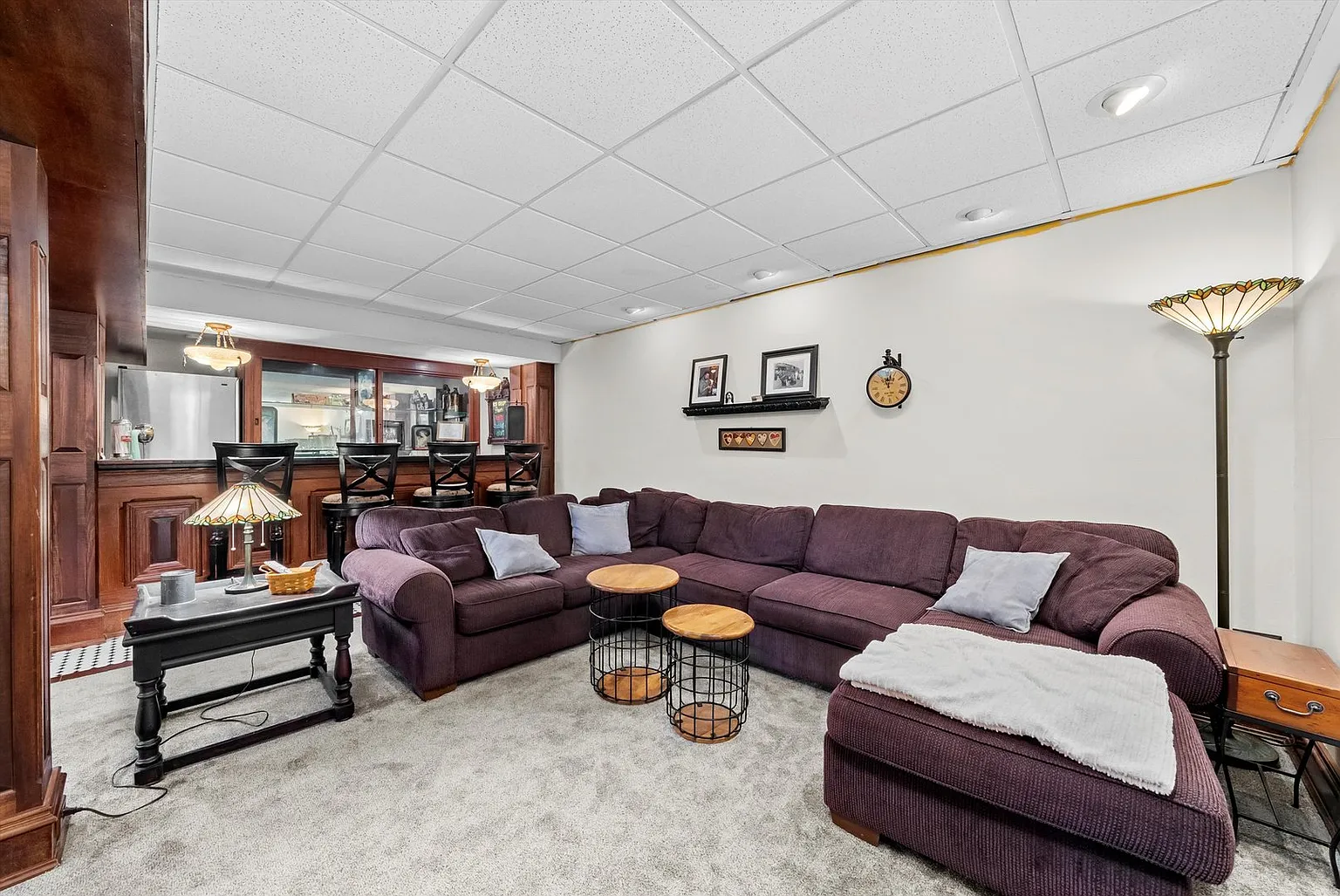 Cozy basement living area with purple sectional sofa, side tables, gray carpet, and bar area.