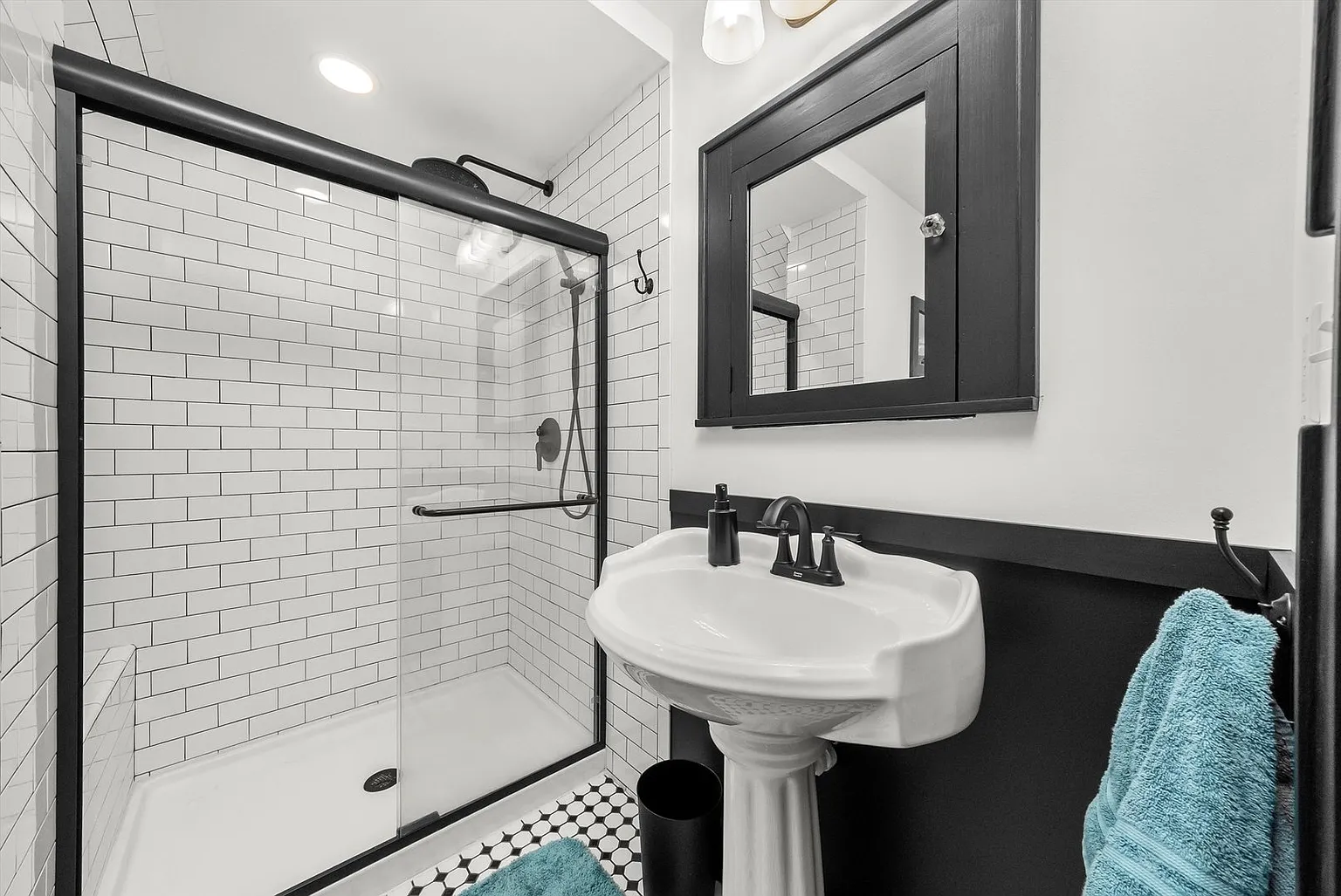 Modern bathroom with glass shower, black frame, white subway tiles, pedestal sink, mirror, and turquoise towel.
