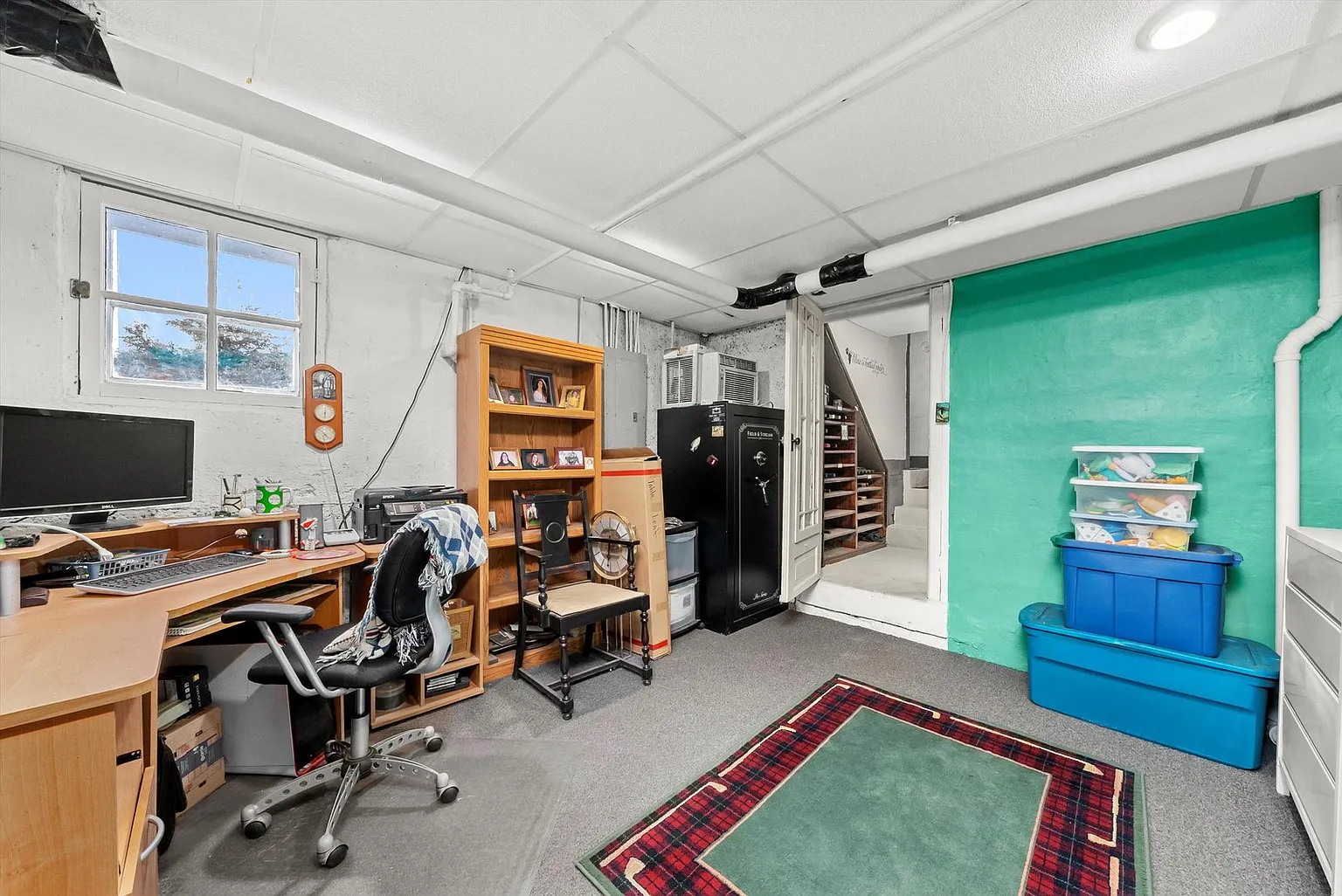 Basement office with desk, computer, chair, bookshelf, mini fridge, stairs, green wall, and rug.
