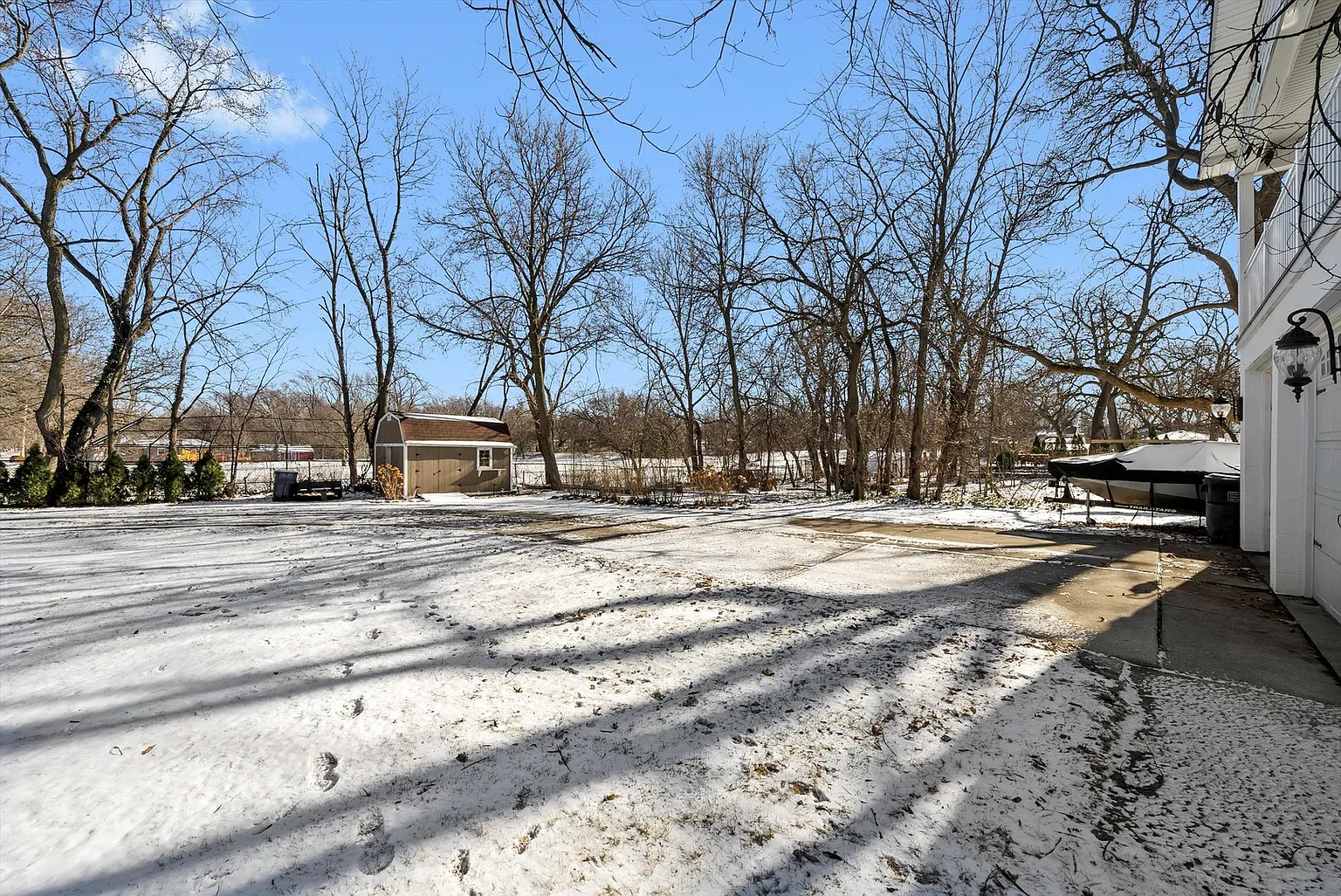 Snow-covered backyard with bare trees, shed, boat, and sunny winter shadows.