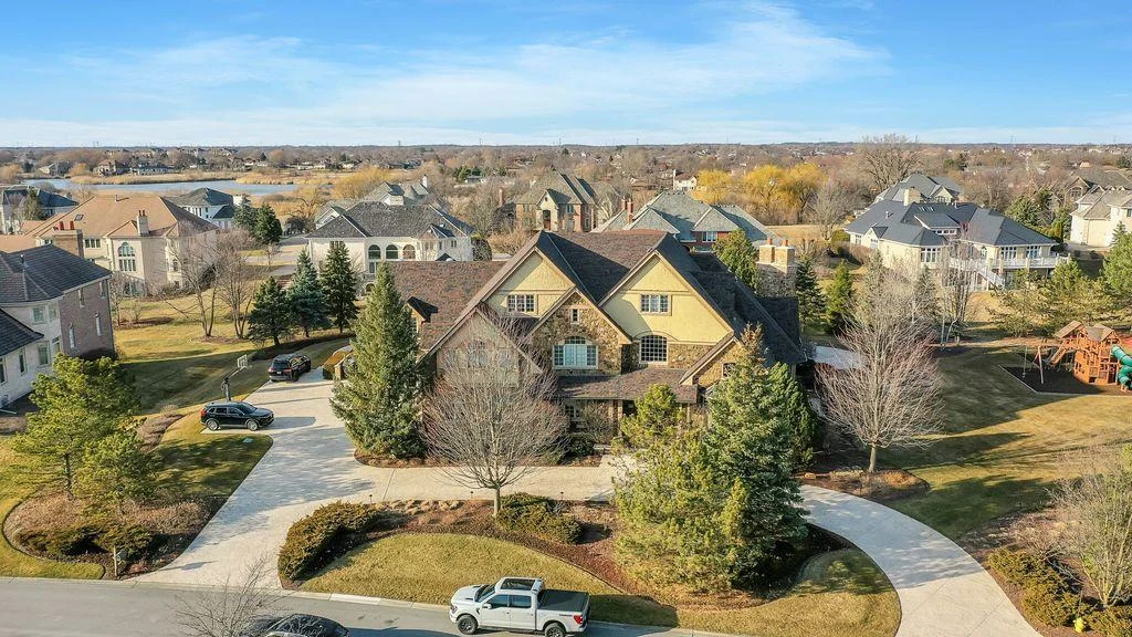 Aerial view of large home with manicured lawn, trees, nearby houses, and blue sky.