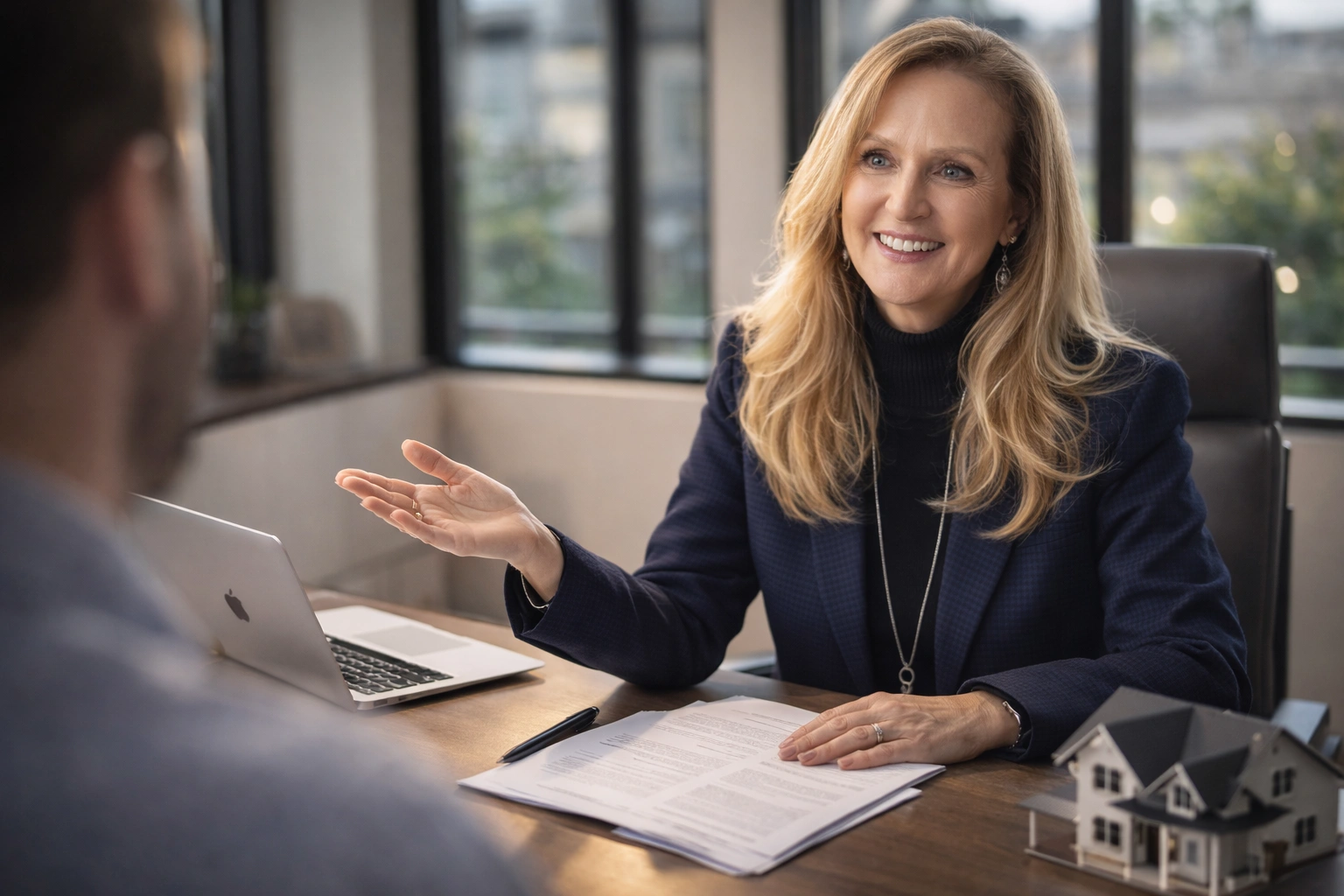 Carla at desk, smiling, wearing navy blazer, with laptop, papers, and model house.