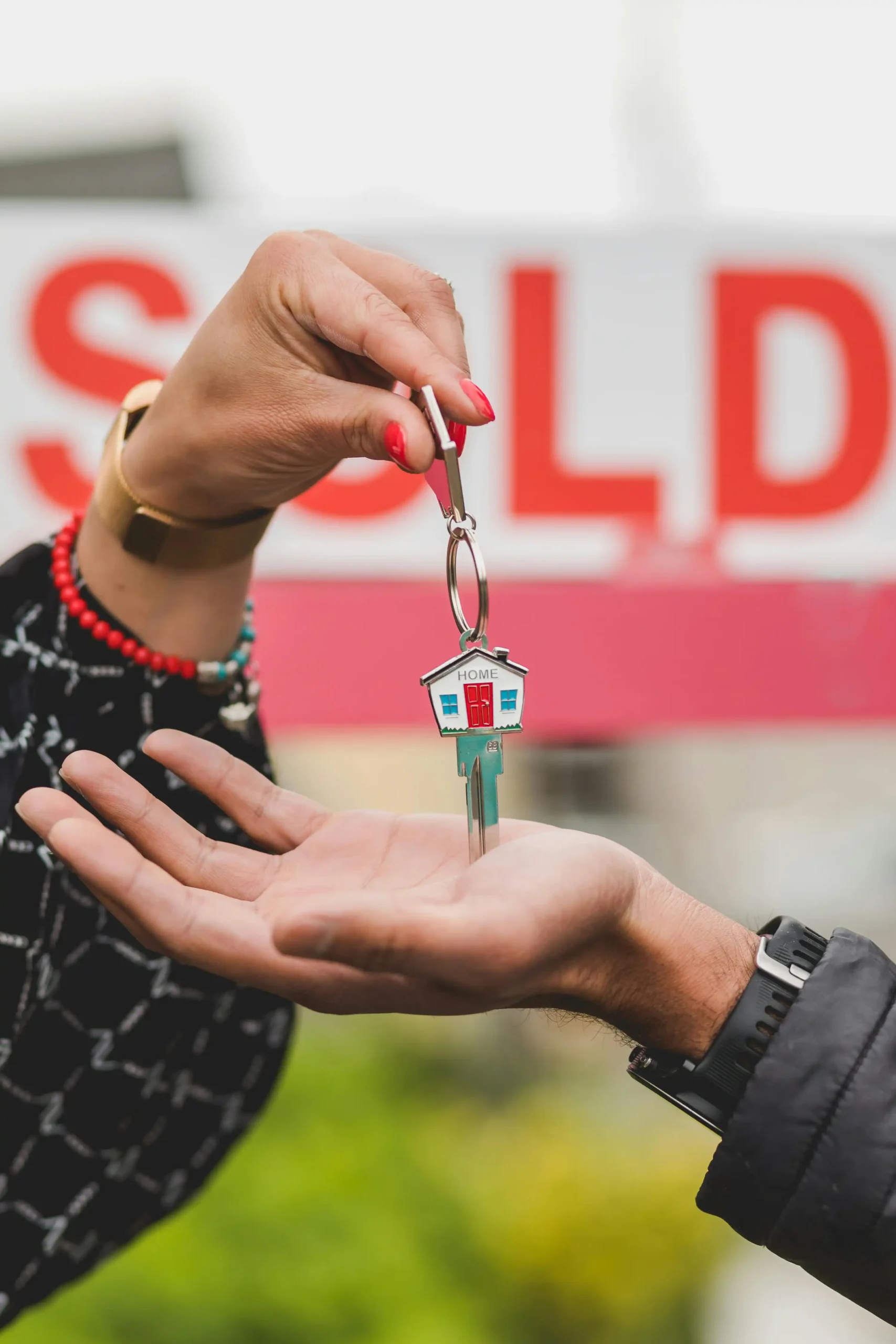 Hand holding house keychain with sold sign in the background.