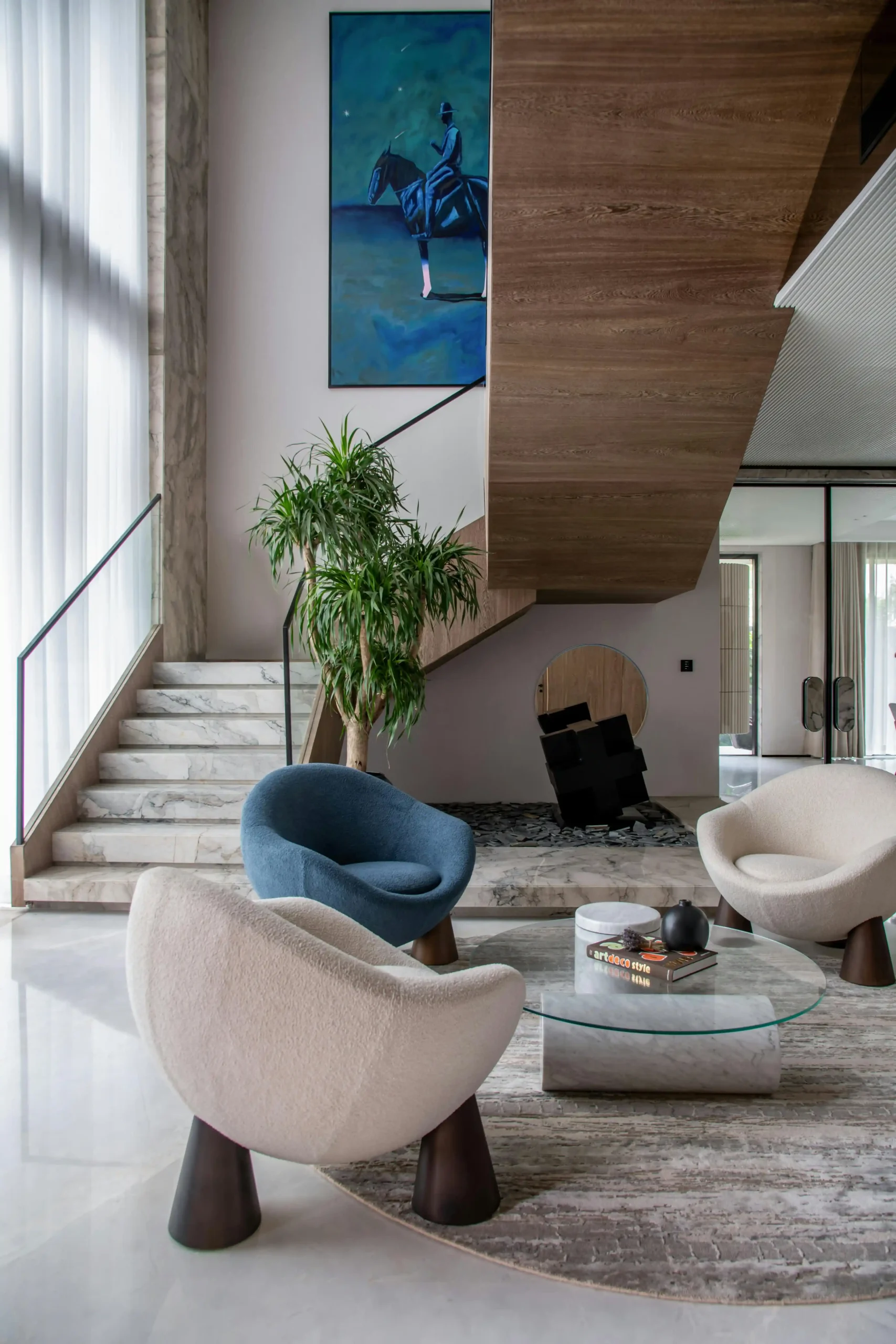 Modern living space with curved chairs, glass coffee table, staircase, and wall art.
