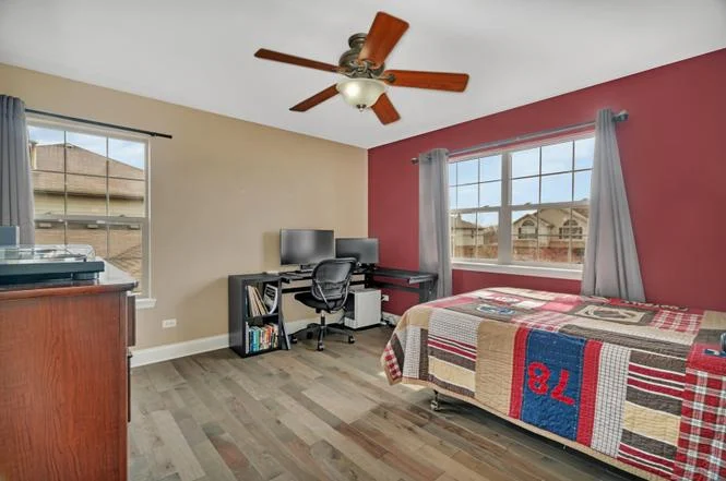 Bright bedroom with colorful quilt, desk setup, ceiling fan, and large windows.