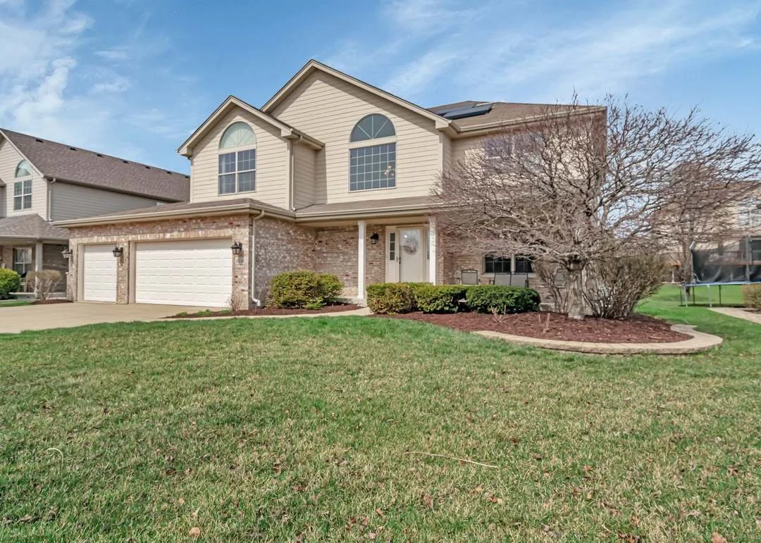 Two-story suburban house with brick facade, three-car garage, lawn, and shrubs.