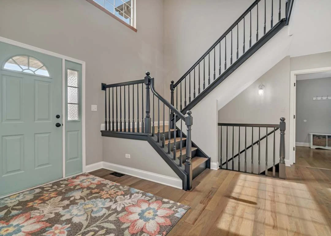 Foyer with light blue front door, staircase with black railings, and decorative rug.